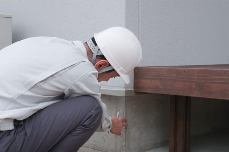 A home inspector wearing a white hard hat examines a house foundation for potential structural issues.